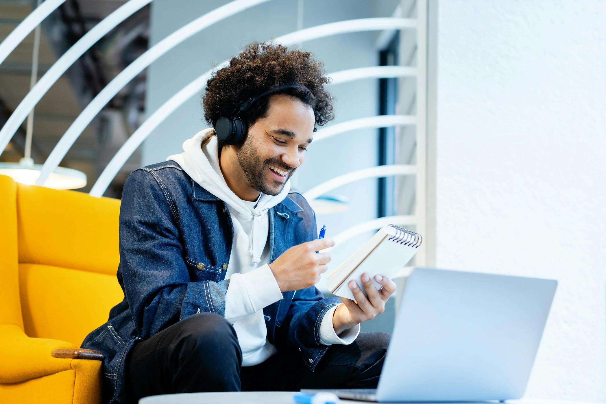 Um freelancer sorridente usando fones de ouvido e uma jaqueta jeans está sentado em uma cadeira amarela, segurando um caderno e uma caneta, olhando para a tela de um laptop em um ambiente moderno e iluminado — talvez explorando uma Calculadora de Faturamento.