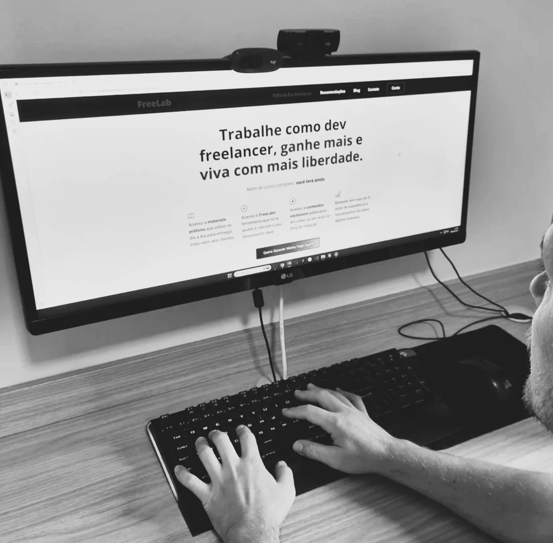 Uma pessoa digita em um teclado em uma mesa, olhando para um monitor widescreen que exibe um site em português sobre trabalho remoto e tendências e previsões de freelancers tecnológicos para 2026.