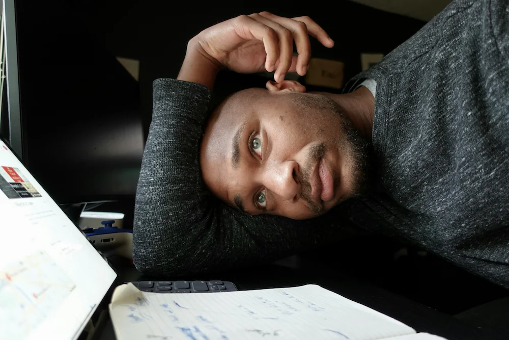 Um freelancer apoia a cabeça no braço em uma mesa, parecendo cansado ou contemplativo. À sua frente, há um laptop aberto, um caderno com anotações escritas à mão e uma calculadora.
